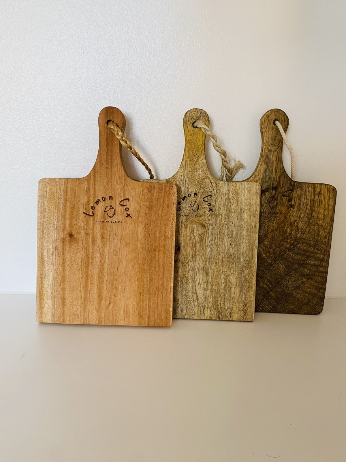 Three rectangular Lemon Cox mango wood serving boards in different wood tones, branded, with rope handles